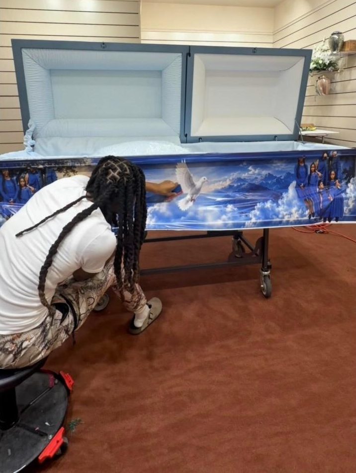 A Suave Wraps installer kneeling beside a casket, smoothing a printed wrap of blue sky, clouds and a white dove