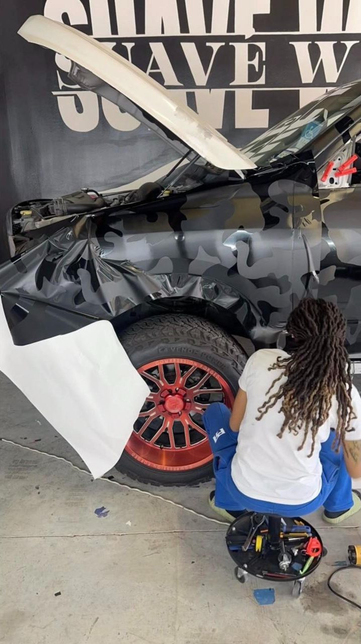 A Suave Wraps installer kneeling on a tool stool, laying camo vinyl around a truck's front wheel